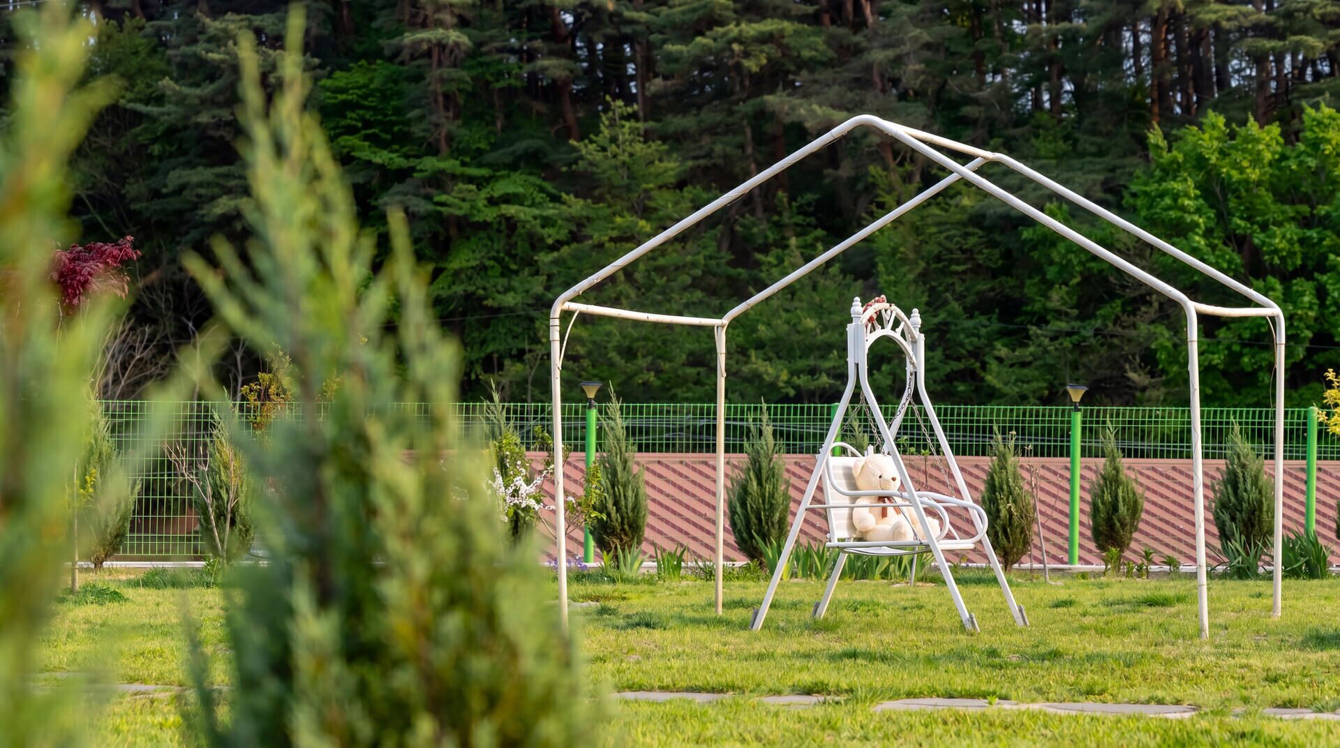 A swing set with a white metal frame and a swing seat, surrounded by a green fence and trees.