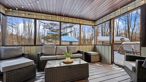 A covered outdoor seating area with wicker furniture, a glass table, and a view of trees and a house.