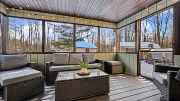 A covered outdoor seating area with wicker furniture, a glass table, and a view of trees and a house.