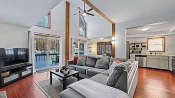 A modern living room with a large flat-screen TV, grey sofas, a coffee table, and a dining area with a kitchen in the background.