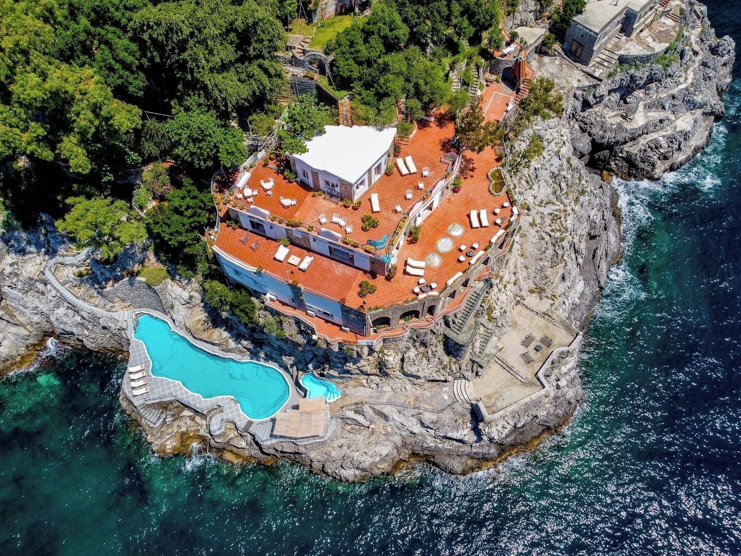 Vista aérea de um hotel à beira de um penhasco, com piscina, área de estar externa e costa rochosa.