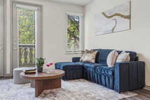 Ein Wohnzimmer mit Ecksofa, rundem Couchtisch und einem großen Fenster mit Jalousien.