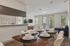 Ein zeitgemäßer Essbereich mit Holztisch, gedeckt für eine Mahlzeit, eine Küche mit Spüle und Schränken sowie ein Wohnbereich mit Sofa und Fenstern.