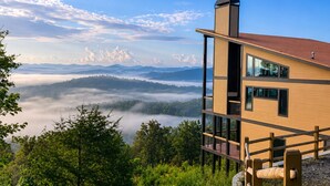 A modern building with a wooden deck, a chimney, and a view of a forested valley and mountains. - Above the Clouds Luxury Retreat with Hot Tub and Epic Views (Morganton)