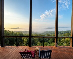Une terrasse en bois avec deux chaises et une petite table, d’où l’on peut admirer un paysage boisé avec des montagnes à l’horizon.