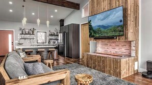 A modern living room with a wooden TV stand, a sofa, and a kitchen in the background. - Above the Clouds Luxury Retreat with Hot Tub and Epic Views (Morganton)
