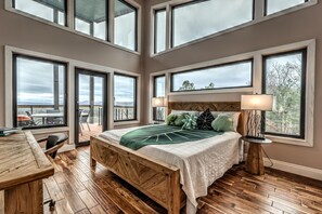 A bedroom with a large bed, multiple windows, a wooden floor, and a view of the outdoors.
