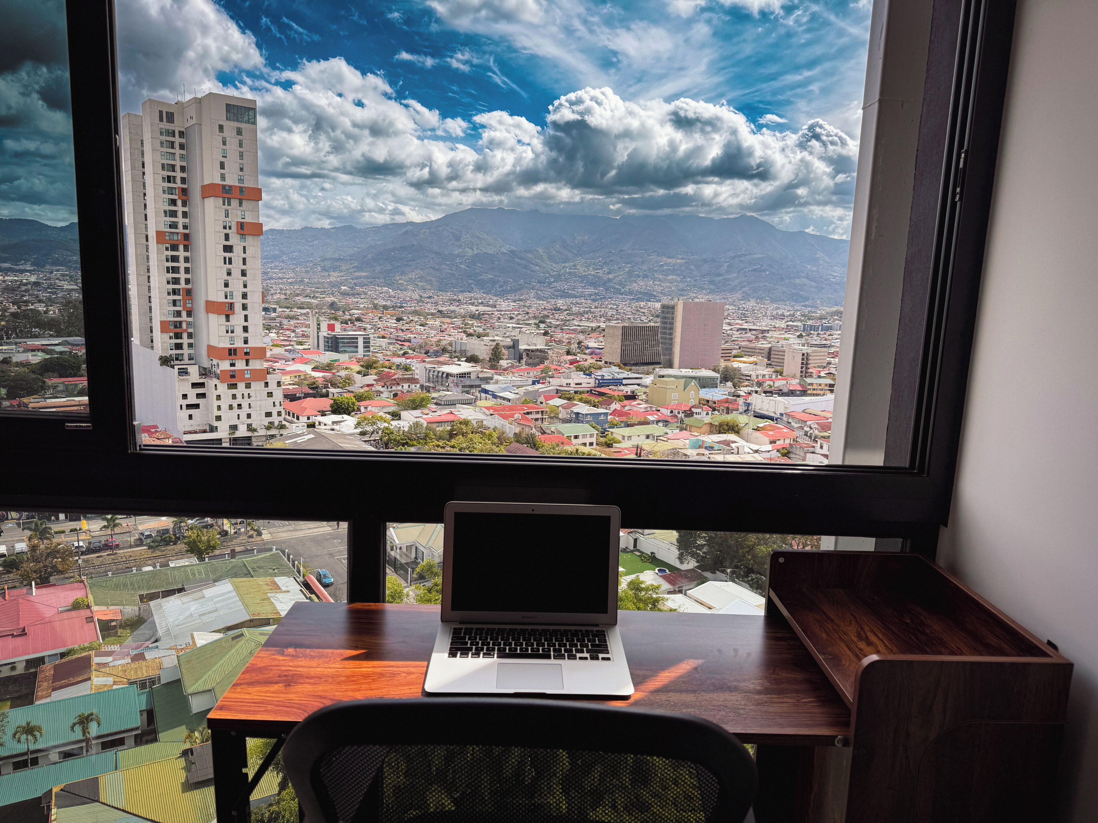 A laptop on a desk with a view of a cityscape through a window.