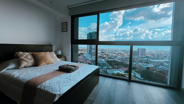 Quarto moderno com uma grande janela, cama com almofadas e vista para o horizonte urbano.