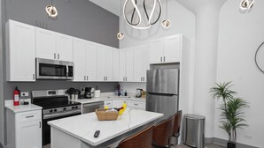 A modern kitchen with white cabinets, stainless steel appliances, a central island with chairs, and a potted plant.