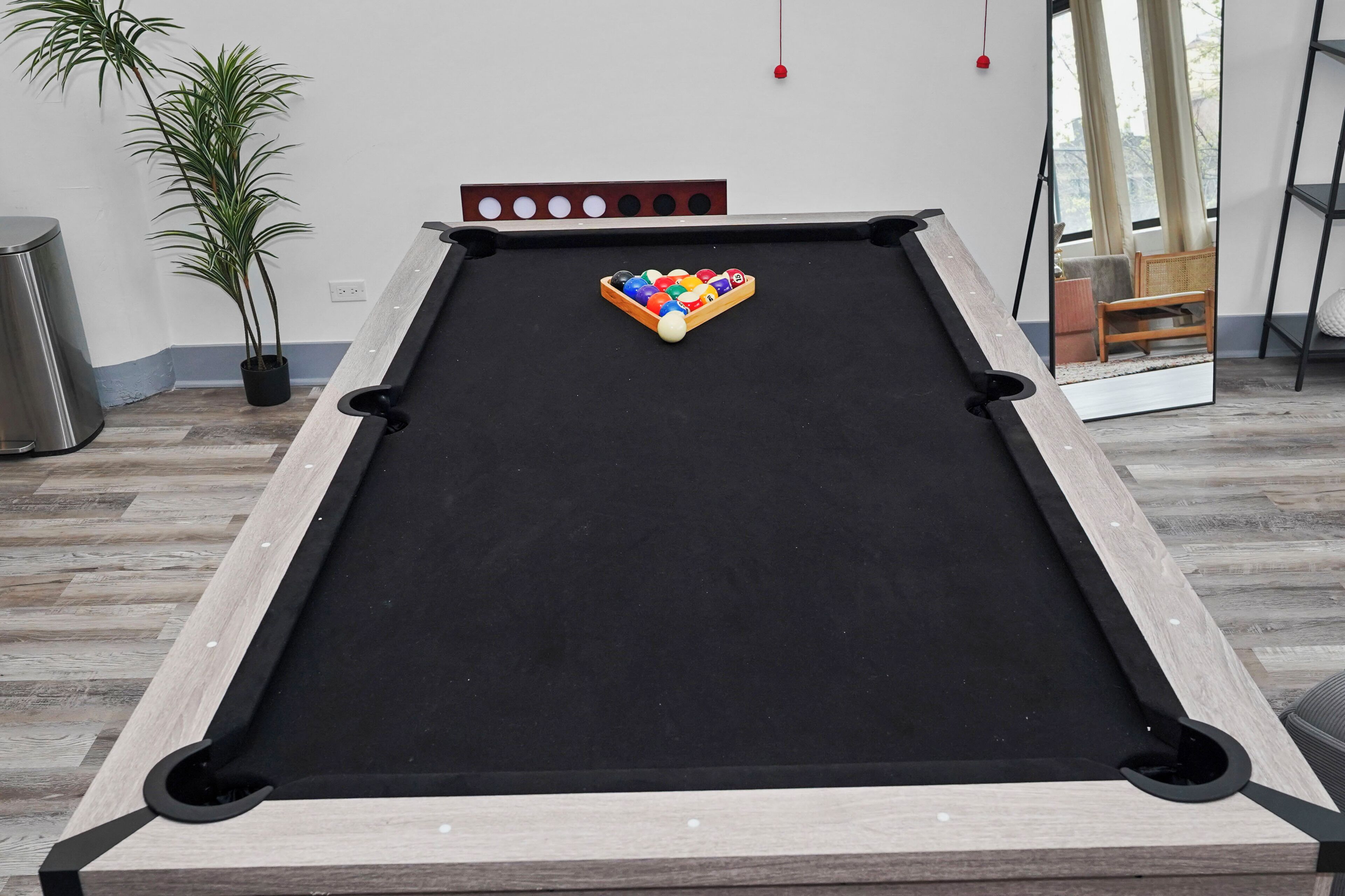 A pool table with a wooden frame, a black felt surface, and a corner pocket. There is a potted plant to the left and a window with curtains in the background.