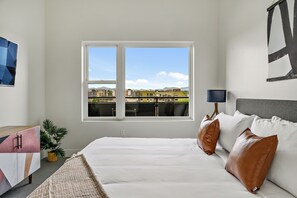 Un dormitorio con un ventanal grande, una cama, una mesita de noche, una lámpara y una planta en maceta.