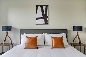 Una cama con sábanas blancas y dos almohadas marrones, flanqueada por dos mesitas de noche con lámparas, en una habitación con cabecero gris y una obra de arte abstracto colgada en la pared.