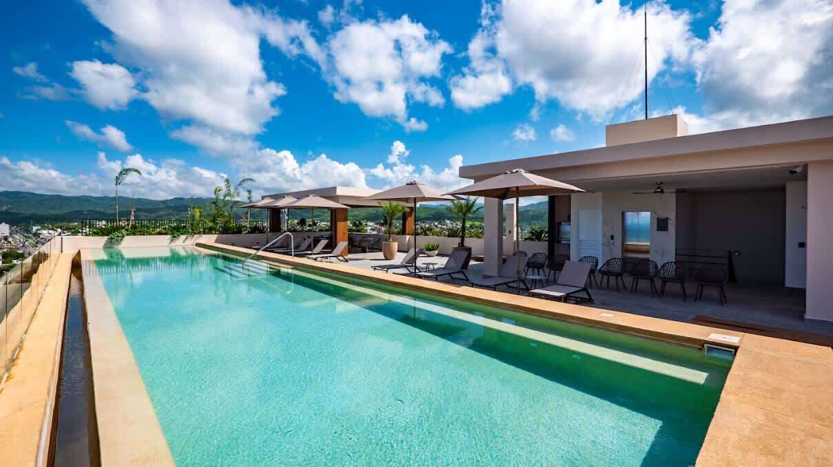 A swimming pool with lounge chairs and umbrellas, surrounded by a modern building with a view of mountains.