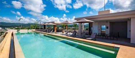 A swimming pool with lounge chairs and umbrellas, surrounded by a modern building with a view of mountains.