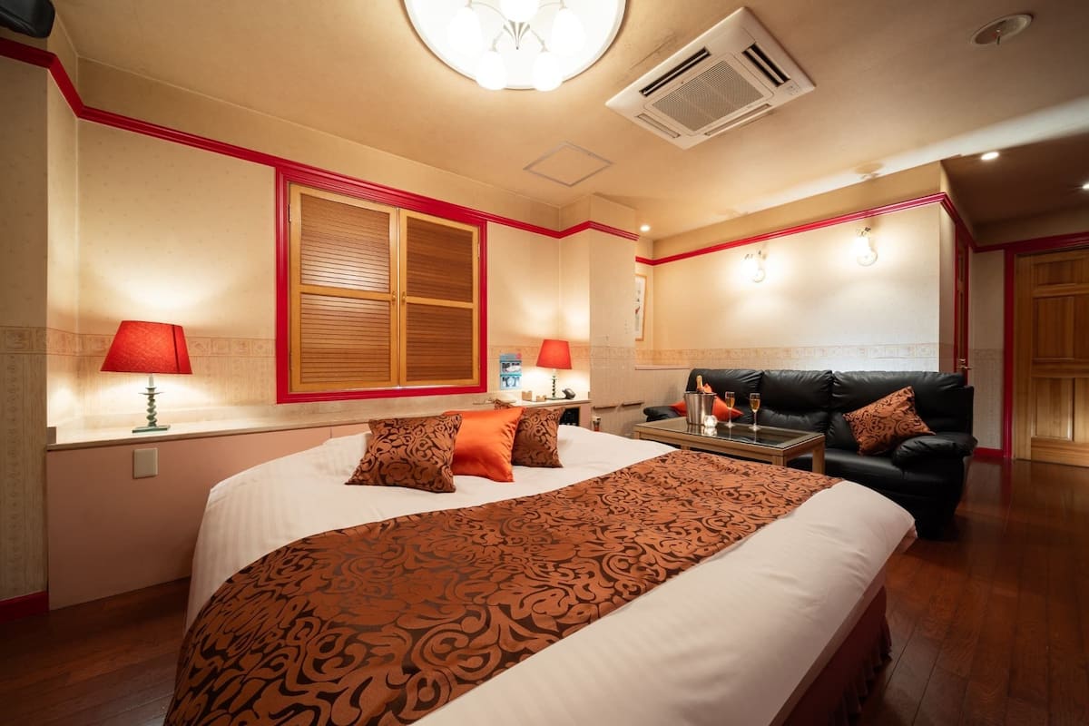 A hotel room with a large bed, a black leather sofa, a small table with a lamp, and a window with wooden shutters.