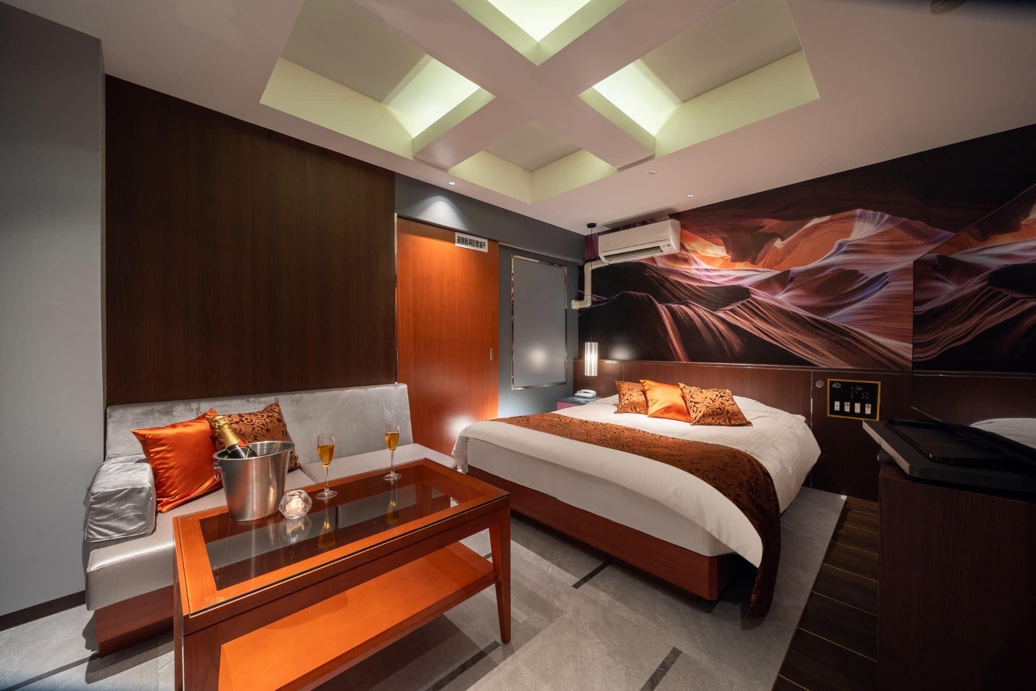 Habitación de hotel con cama, sofá, mesita de vidrio con una botella y un vaso, y aire acondicionado instalado en la pared.
