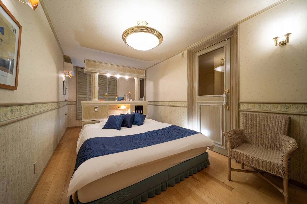 A bedroom with a bed, a chair, a vanity with lights, a mirror, and a door.