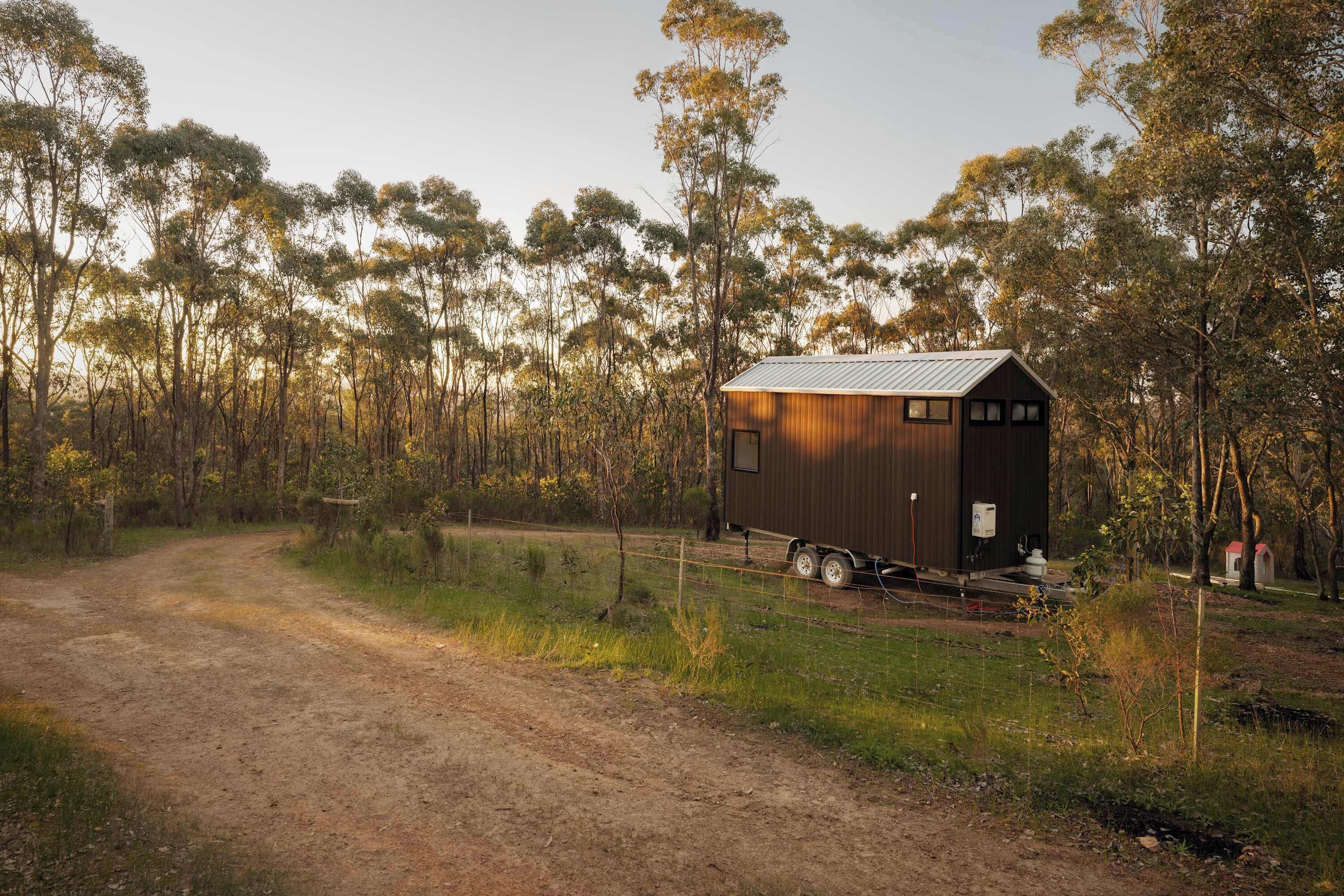 A small, single-story wooden structure with a metal roof, surrounded by a forested area with a dirt path leading to it.