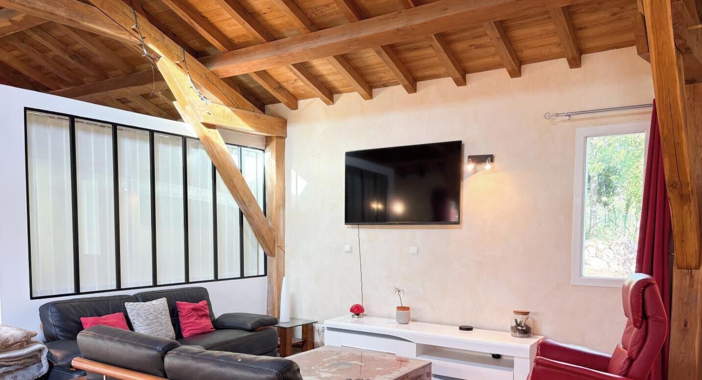 A modern living room with a wooden ceiling, a flat-screen TV mounted on the wall, a sofa with cushions, and a window with curtains.