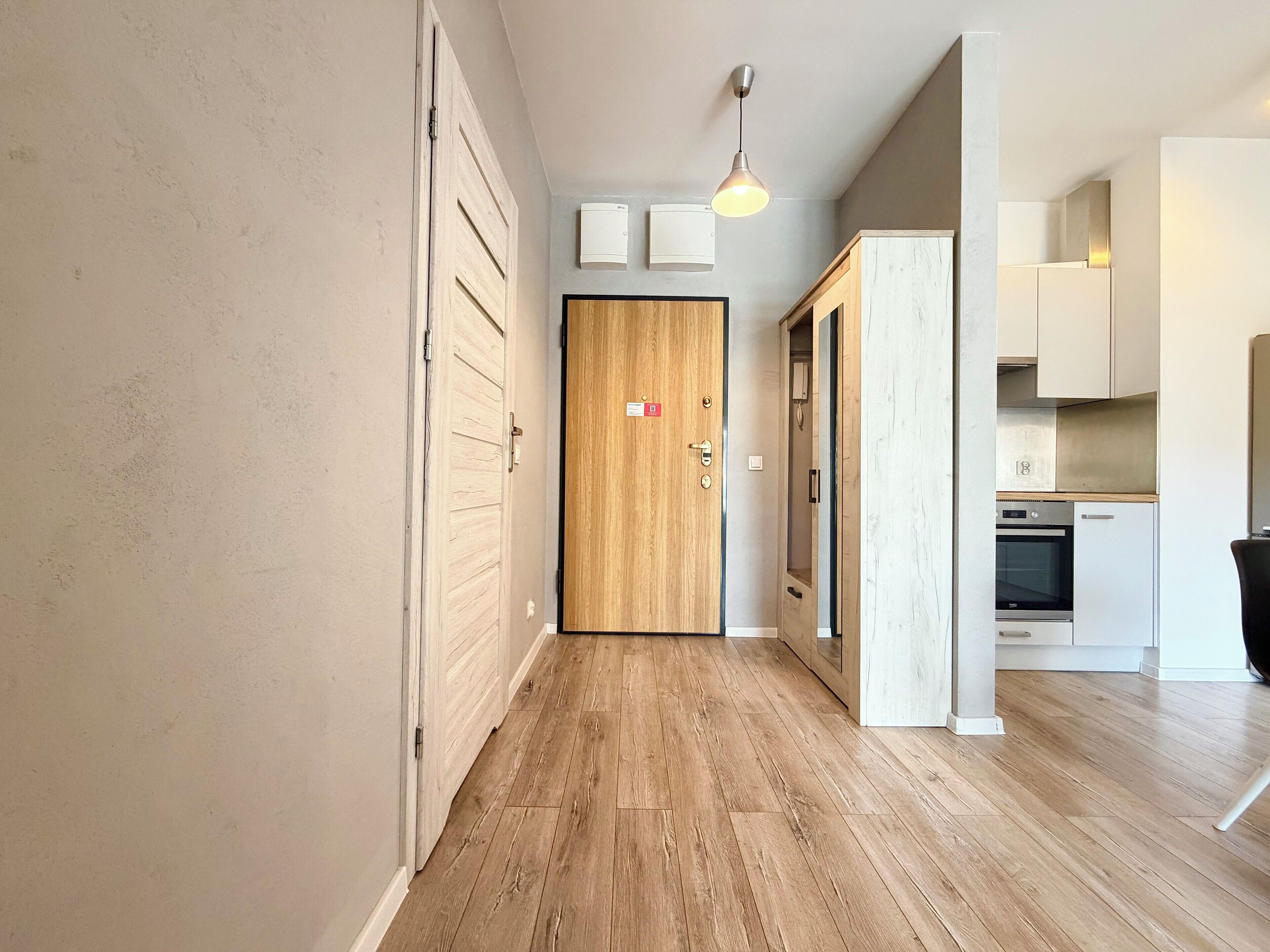 A hallway with wooden flooring, a wooden door, and a kitchen area with white cabinets and appliances.