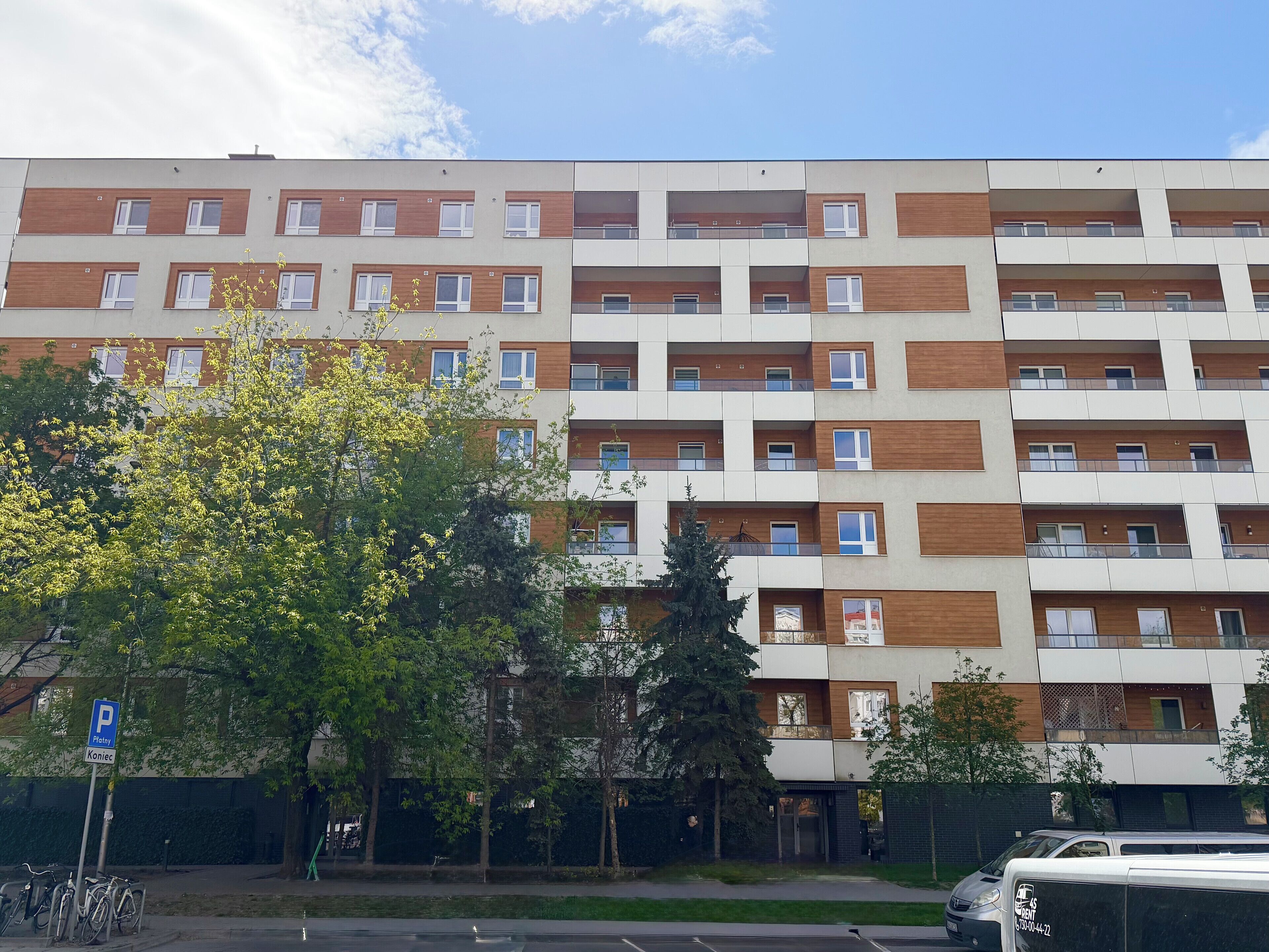 A multi-story residential building with balconies, a parking area, and a tree in the foreground.