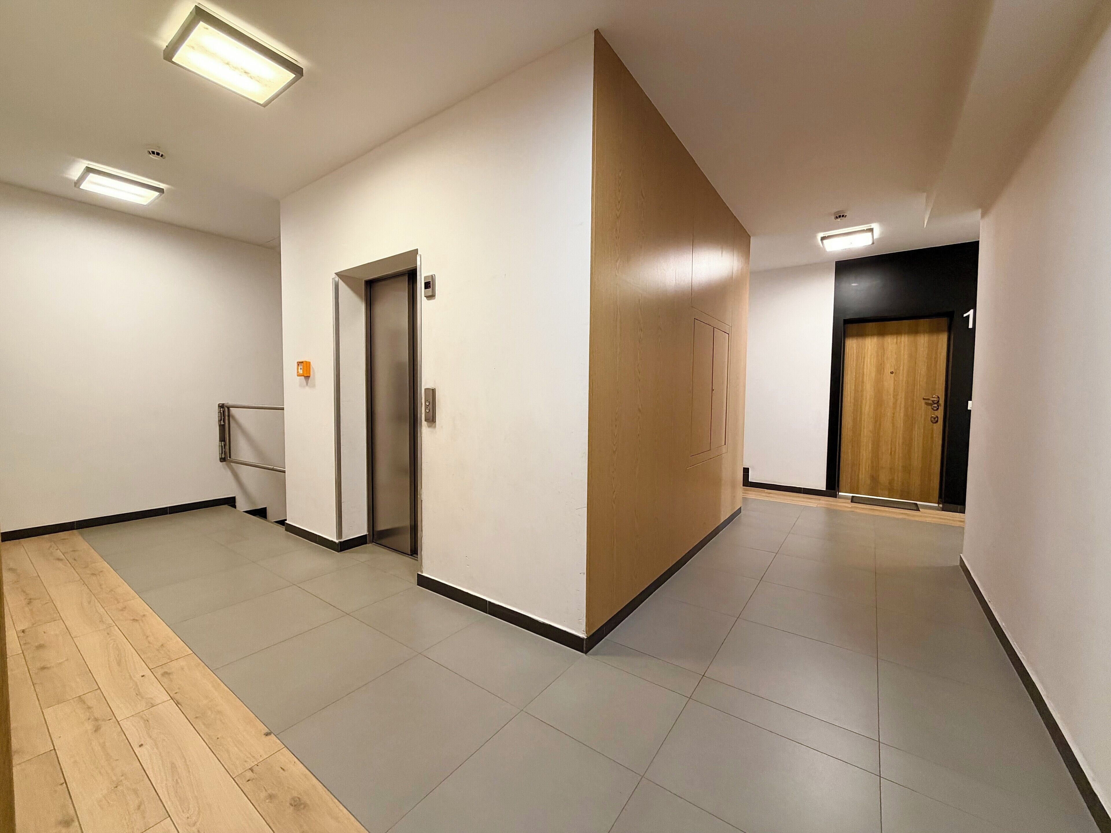 A hallway with a wooden floor, a door with a keypad, and a wall-mounted light fixture.