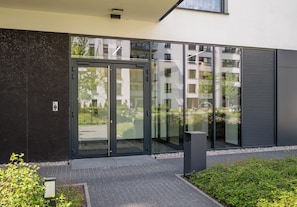 Ein zeitgemäßes Gebäude mit Eingang, Glasüren, einer kleinen Außenanlage mit Pflanzen und einem gepflasterten Weg zum Eingang.