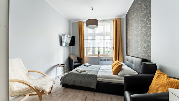 Une chambre moderne avec un lit, deux fauteuils, un téléviseur et une fenêtre avec des rideaux.