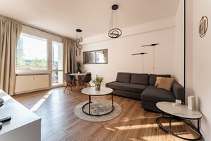 Ein modernes Wohnzimmer mit einer Couch, einem runden Couchtisch, einem Esstisch mit Stühlen und einem großen Fenster mit Vorhängen.