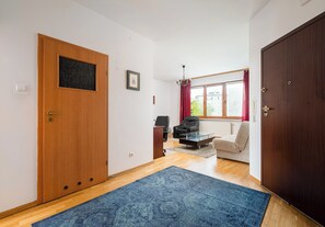 A room with a wooden door, a blue rug, a glass coffee table, a beige sofa, and a window with red curtains. - Imielin Homely Apartment (Warszawa)