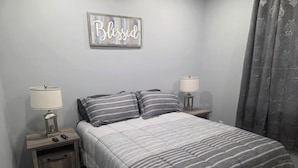 Een slaapkamer met een bed, twee nachtkastjes met lampen, een bordje met de tekst 'Blessed' en een grijs gordijn.
