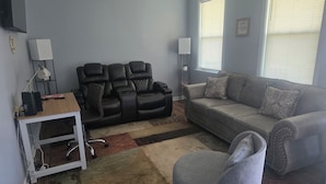 Een woonkamer met een grijze bank, een zwarte leren fauteuil, een wit bureau met een lamp en een klein vloerkleed.