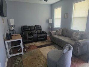 Ein Wohnzimmer mit einem grauen Sofa, einem schwarzen Ledersessel, einem weißen Schreibtisch mit Lampe und einem kleinen Teppich.
