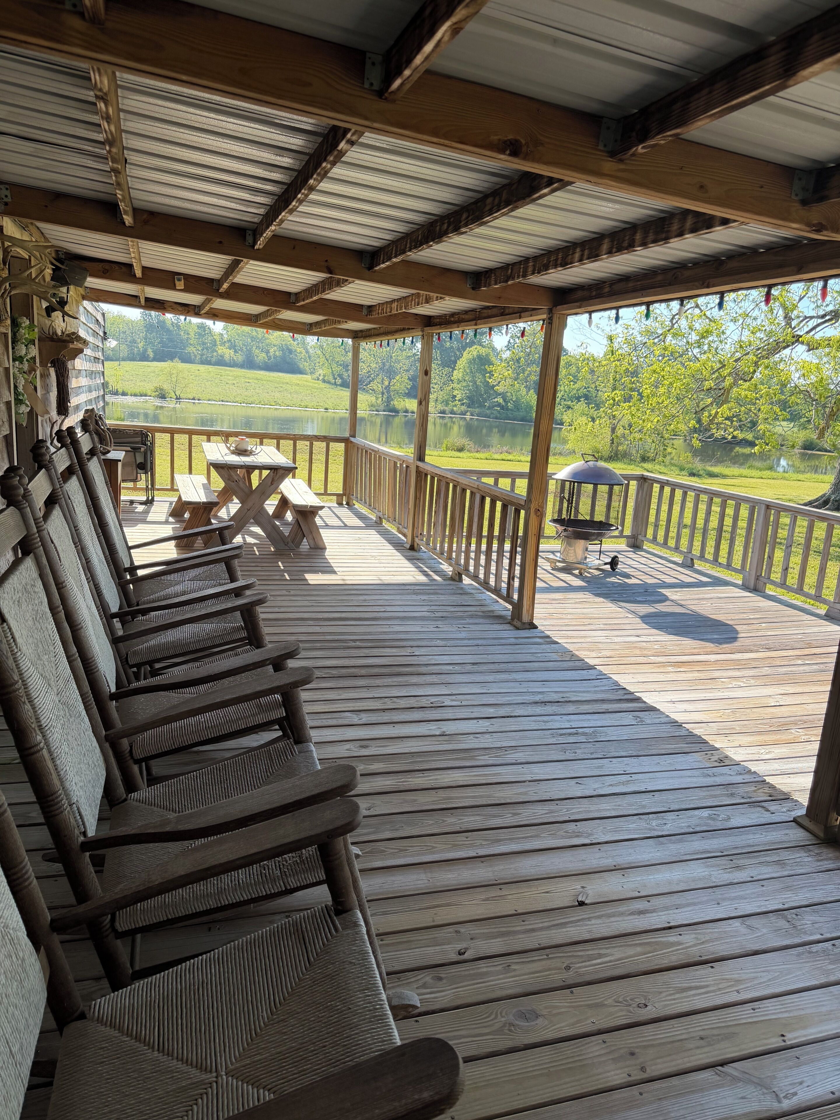 A wooden deck with benches, a table, and a grill, overlooking a lake and greenery.