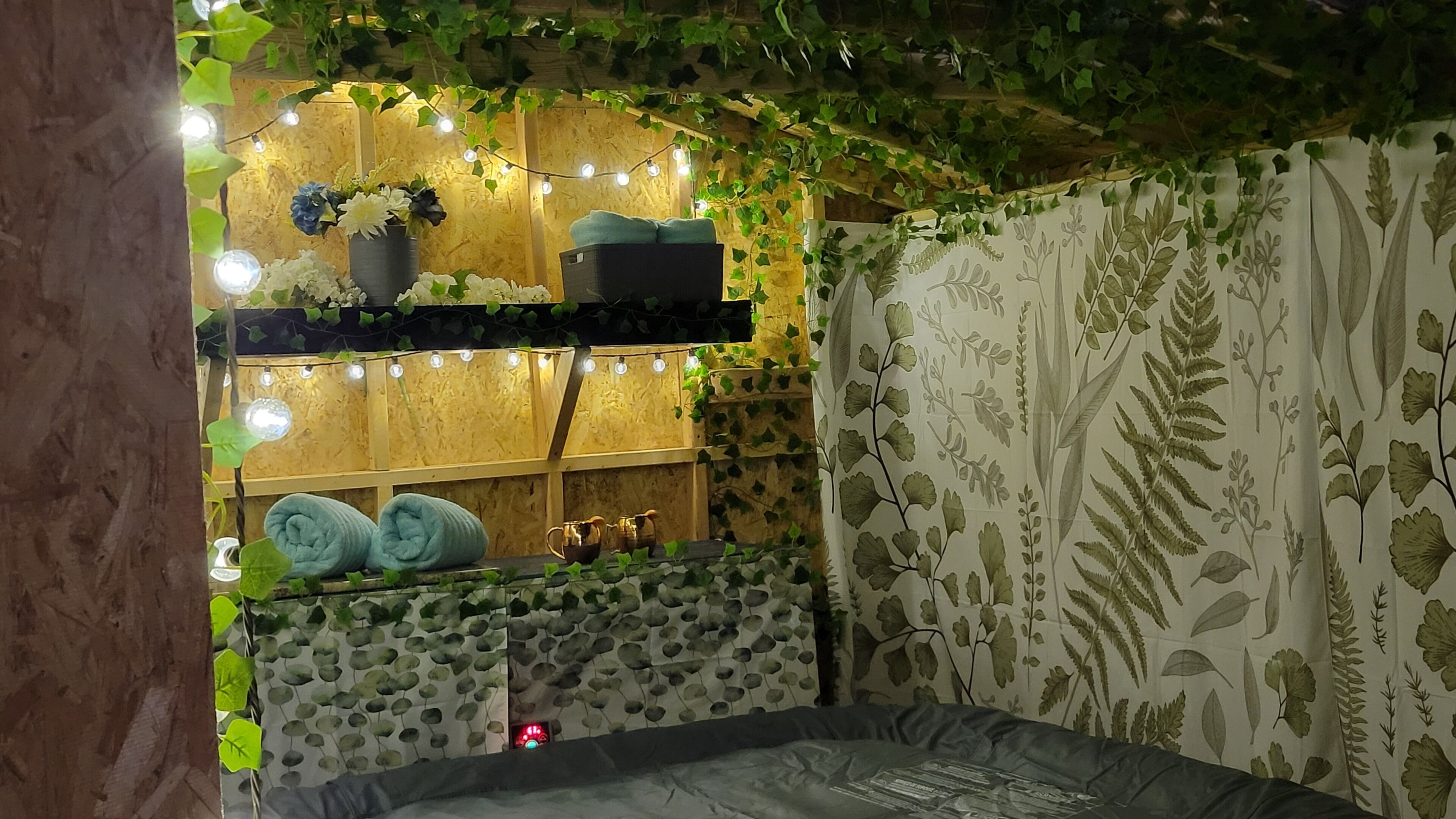 A cozy room with a wooden wall, string lights, a shelf with flowers, a bench, and a patterned curtain.