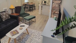 A living room with a sofa, a coffee table, and a TV stand. There is a dining area with a table and chairs. The room has a stone accent wall and a window with curtains. - The Cozy Cottage at the base of Pikes Peak with private hot tub (Cascade-Chipita Park)