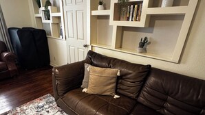 Un canapé en cuir marron, un tapis à motifs, une porte blanche et des étagères intégrées ornées de plantes et de livres.