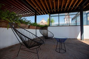 A covered outdoor area with two black wireframe chairs and a small round table, a brick floor, and a view of a building through large windows. - La Casona de Puebla | 2bd Apt | Centro Histórico (Puebla)