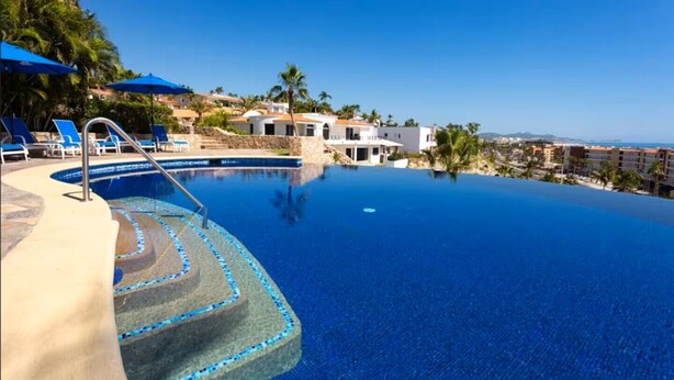 A clear blue infinity pool with steps leading into it, surrounded by lounge chairs and umbrellas, with a view of a coastal area and buildings in the background. - Cozy Condo/Ocean view/ Private balcony/Paseo la Jolla/ San Jose del Cabo (San José del Cabo)