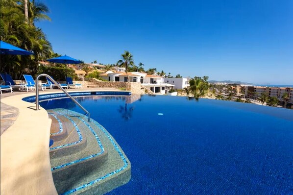 Ein klarer, blauer Infinity-Pool mit Stufen, umgeben von Liegestühlen und Sonnenschirmen, mit Blick auf eine Küstenlandschaft und Gebäude im Hintergrund.