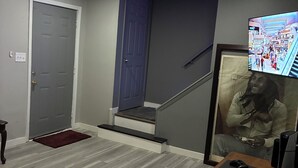 Una habitación con una puerta gris, una escalera, un cuadro enmarcado y un televisor mostrando un centro comercial.