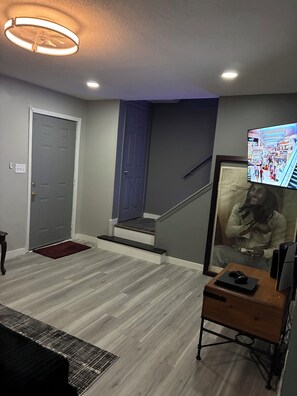 Ein Zimmer mit einer grauen Tür, einer Treppe, einem gerahmten Bild und einem Fernseher, der eine Einkaufspassage zeigt.
