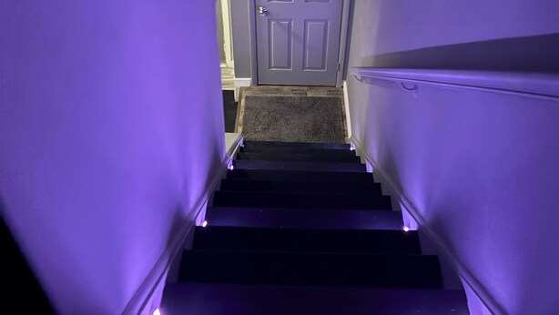 Una escalera estrecha con una puerta al final, escalones alfombrados e iluminación morada.
