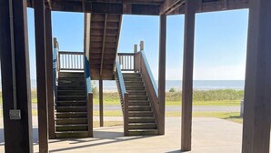 A covered outdoor area with two wooden staircases leading to a higher level, a concrete floor, and a view of the landscape. - Beachfront • Sleeps 16 • King Bed • 6 Queens (Bolivar Peninsula)