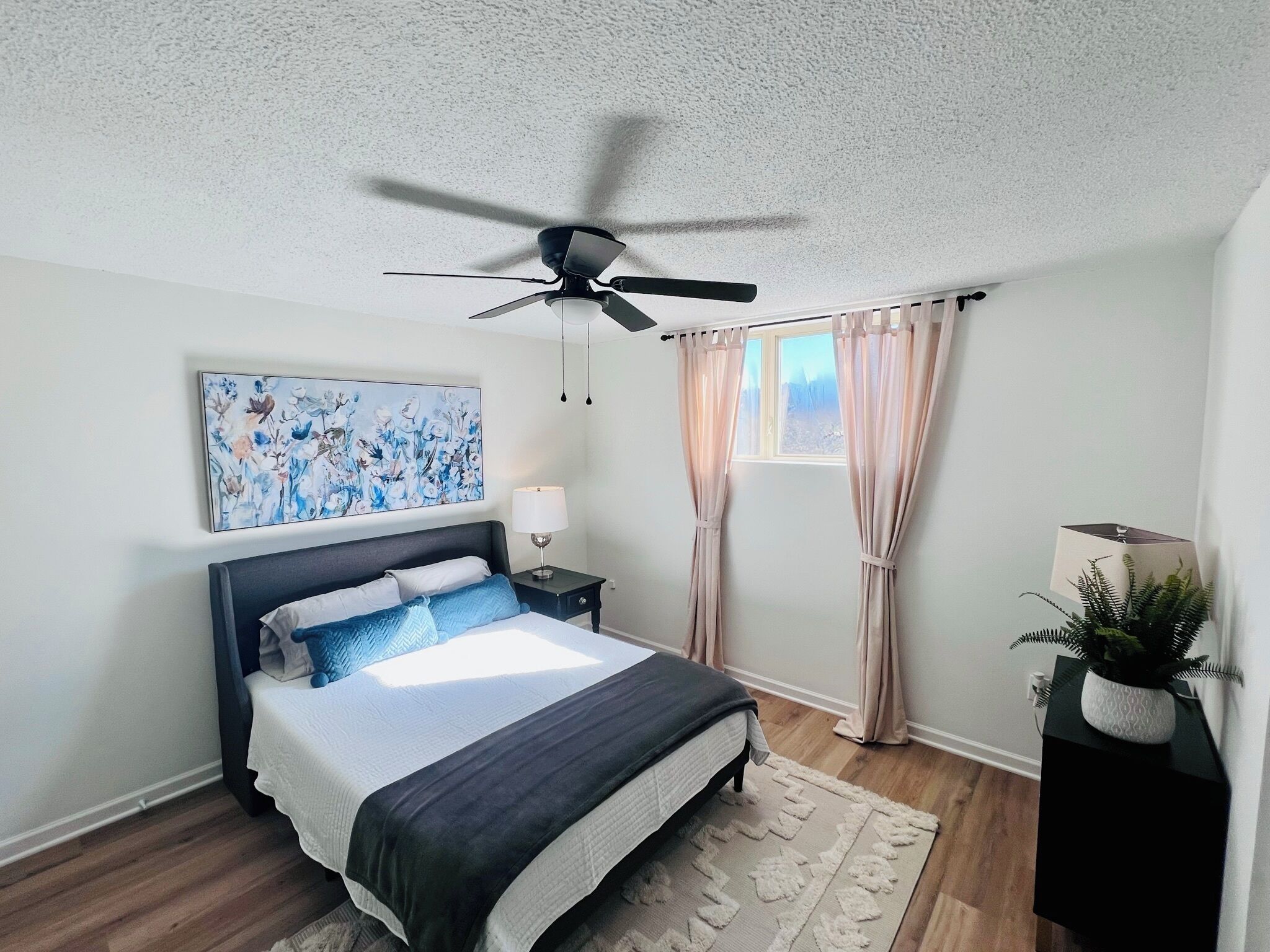 A bedroom with a bed, a ceiling fan, a window with curtains, a nightstand with a lamp, and a wall-mounted painting.