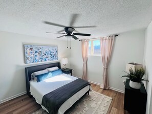Ein Schlafzimmer mit Bett, Deckenventilator, Fenster mit Vorhängen, Nachttisch mit Lampe und einem an der Wand hängenden Bild.