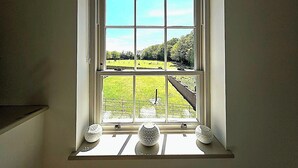 Una ventana con vista a un campo verde y árboles, un marco blanco y dos jarrones decorativos en el alféizar.