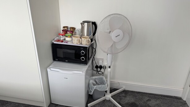 Una cuineta compacta amb microones, nevera petita, cafetera i ventilador portàtil.
