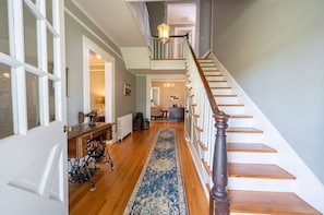 A hallway with a wooden floor, a blue patterned rug, a staircase with wooden steps and a dark wooden handrail, and a white door on the left.